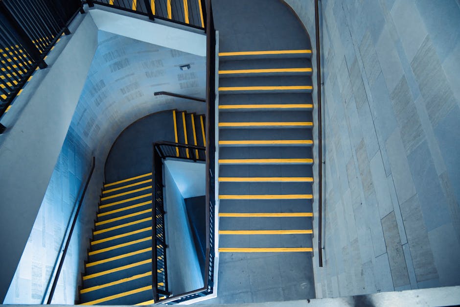 A view looking upwards through a multi-storey staircase of a residential property, showing black metal railings and painted white walls. The staircase features a geometric, spiral design with landings