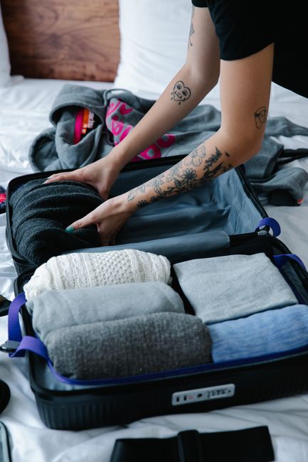 A person packing a black suitcase on a bed with white and gray bedding, surrounded by clothing items such as folded sweaters, socks, and undergarments. The individual, with visible tattoos on their ar