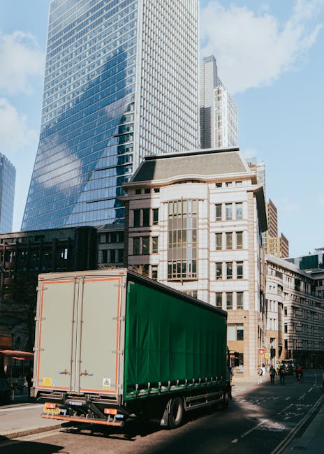 A large green and beige moving truck parked on a city street in front of tall modern office buildings and historic multi-storey structures during daylight. The truck is positioned near the curb, with 