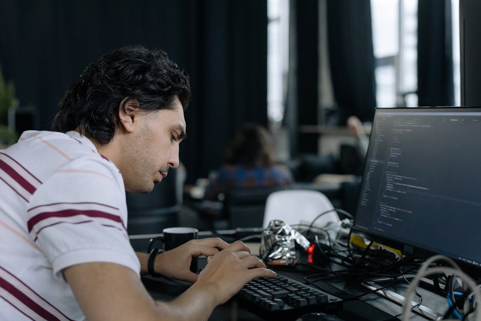 A close-up of a person's hand operating a silver laptop placed on a wooden desk, with the screen displaying lines of code in a text editor. The individual is wearing a black wristwatch and is using th